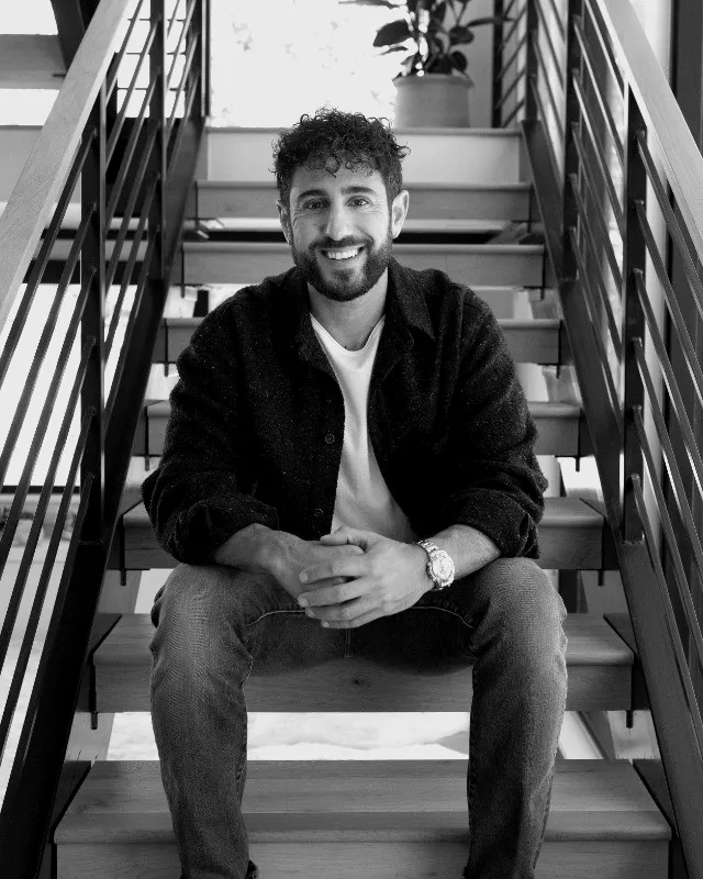 Justin Rezvani sitting on a wooden staircase, smiling, in a dark overshirt and jeans
